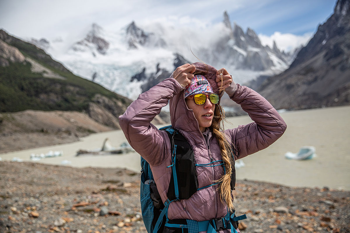 A woman pulling a jacket hood over her head while hiking in a mountainous landscape