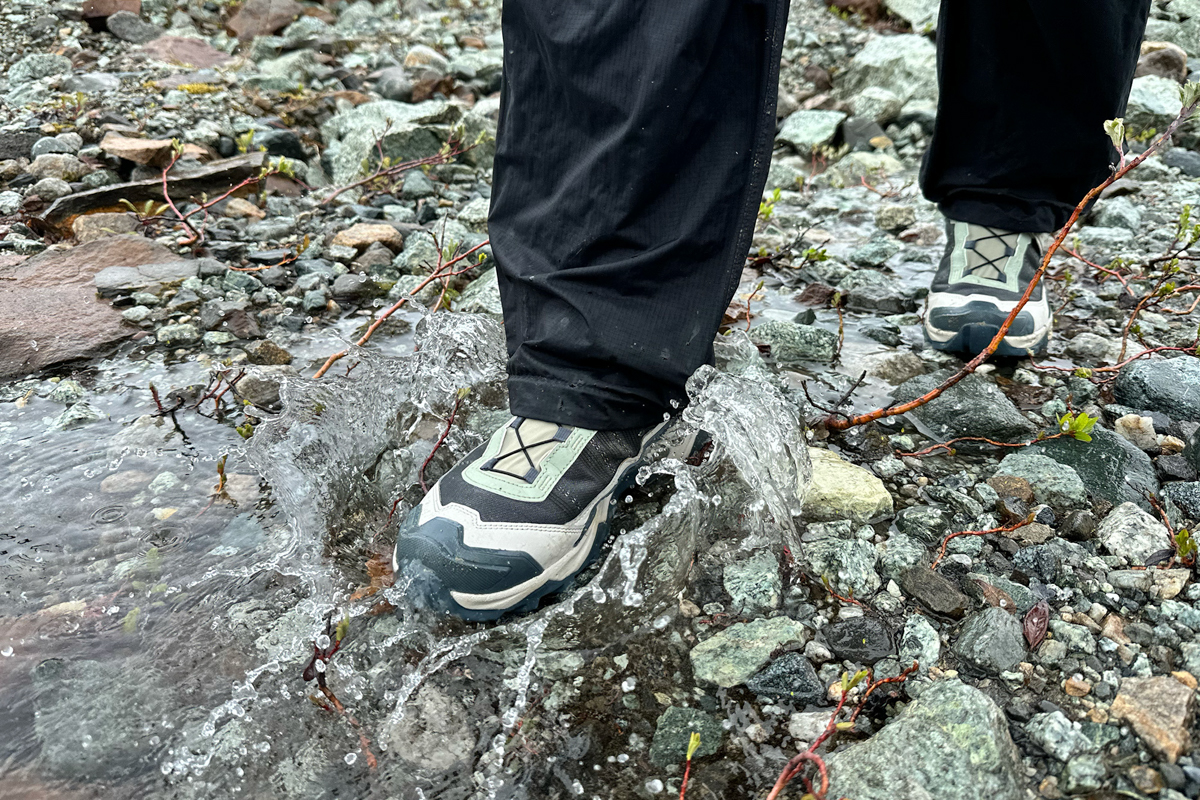 A green and brown hiking shoe stepping into a shallow rocky stream with a splash