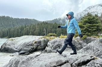 A woman wearing rain boots and a rain jacket walks along rocks on a cloudy day